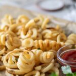A close up of air fried curly fries next to a container of ketchup.