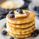 5 pancakes on a white plate with blueberries, butter, and syrup.