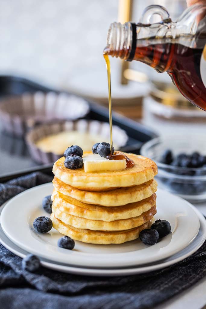 A stack of pancakes with butter and blueberries, as well as syrup being poured overtop.
